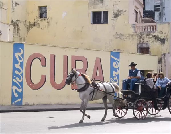 Tourists in a horse carriage passing a wall sign saying Live free Cuba