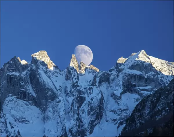 The moon appears behind the snowy mountains illuminating the peaks at sunset, Bondasca Valley