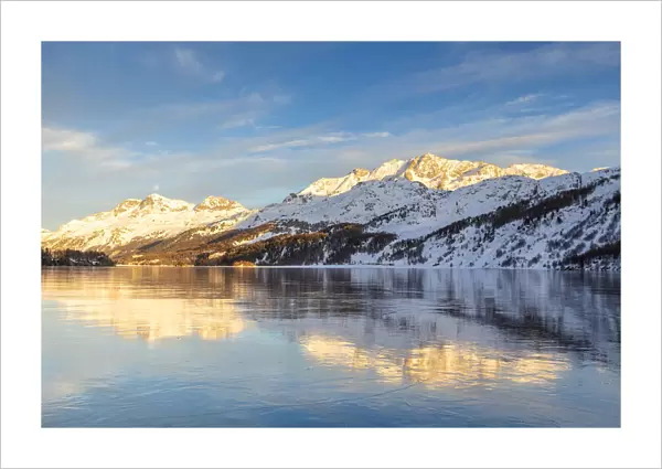 Mountains illuminated by sun at sunset reflected on the icy surfaces of Lake Sils