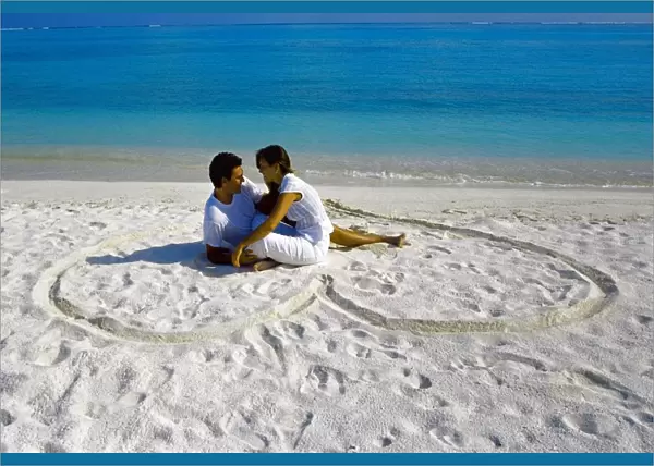 Young couple on beach sitting in a heart shaped imprint on the sand, Maldives