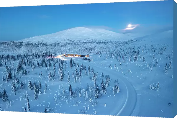 Aerial view of a snowy mountain road in the frozen forest nearby a ski area at dusk, Yllastunturi National Park, Lapland, Finland