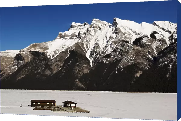 A man walks over a frozen Lake Minnewanka to a boathouse in front of the Canadian