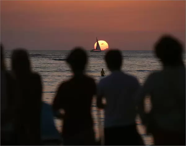 People watch a boat sail in front of the setting sun on New Years Eve on Waikiki Beach