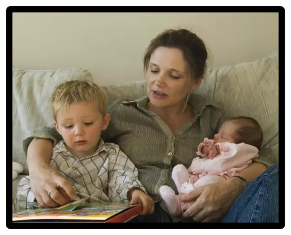 Mother, toddler and new born girl reading childrens book on sofa at home Norfolk UK