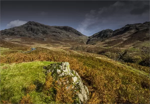 View of road snaking towards upland pass, with volcanic rock in foreground, Hardknott Pass, Lake District N. P