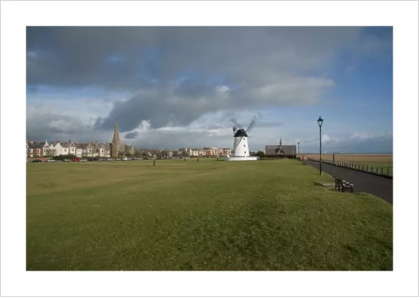 View of grass promenade in seaside resort town, with windmill and old lifeboat house in distance, Lytham Windmill
