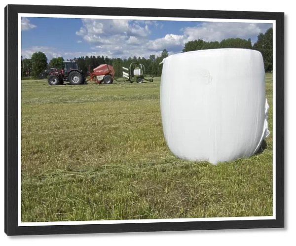 Plastic wrapped round silage bale in field, tractor with baler and mechanical bale-wrapper, in background, Sweden