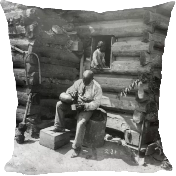 LOG CABIN, c1895. An African American man seated on a tree stump, outside a log