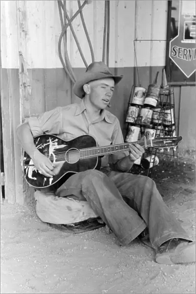 GUITAR PLAYER, 1940. A farm boy playing guitar outside of a filling station in Pie Town