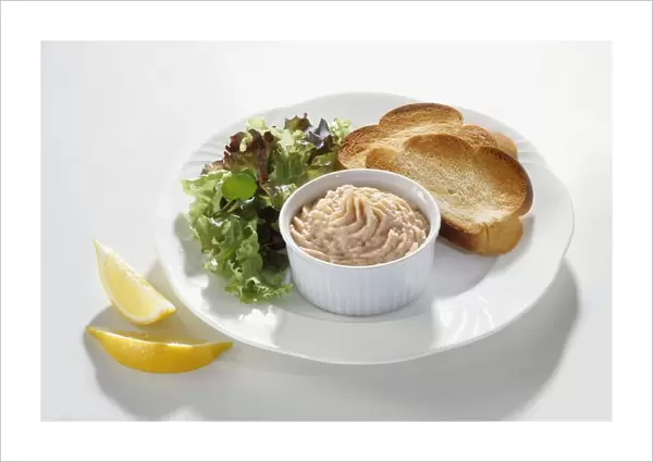 Smoked trout pate in ramekin served on white plate with toasted chollah and lettuce leaves and sliced lemon