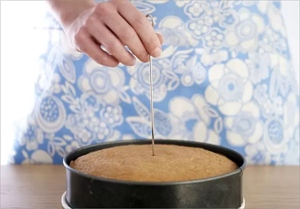Hand inserting metal skewer into baked cake in round cake tin, close-up