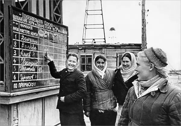 A crew of young women workers, headed by mari volkova (right), have just finished their shift and are reading the results of the high output competition, volkova and her girlfriends are in second place, the heading of the exhibition board reads fulfillment quota for oil output from, , , , 1940s