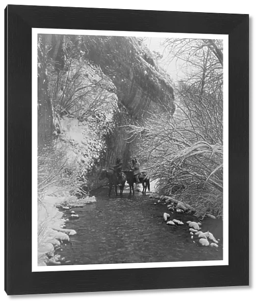 Two Crow Indians on horseback in shallow stream flanked by snow-covered trees, under rock cliff