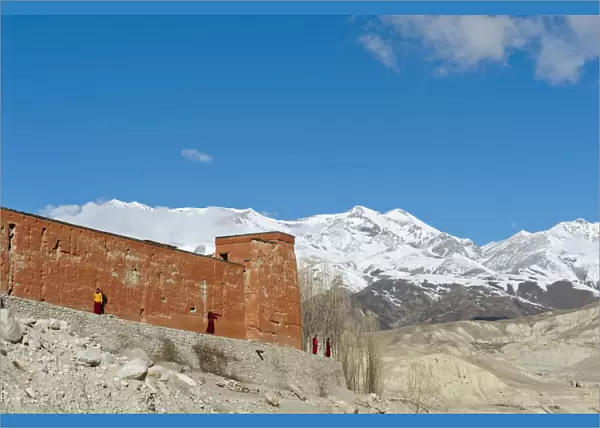 Red wall of a building with monks, Choede Gompa monastery, snow-capped mountains of Mustang Himal mountain range at back, Lo Manthang, Upper Mustang, Lo, Nepal