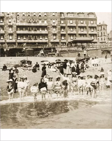 Historic image in sepia of a group of children standing in a row in shallow water with their skirts and knickers pulled up, adults sitting on the beach; Brighton, England
