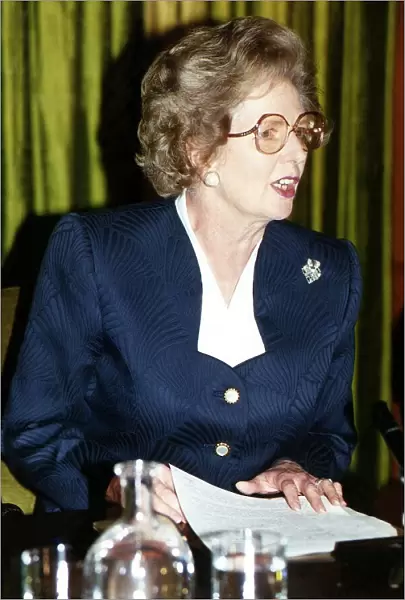 Lady Margaret Thatcher sitting at a table at a press conference wearing glasses