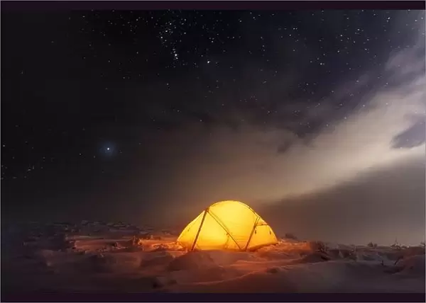 Yellow tent lighted from the inside against the backdrop of glowing city lights and incredible starry sky. Amazing night landscape