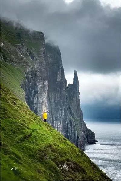 Tourist in yellow jacket looks at Witches Finger cliffs from Trollkonufingur viewpoint. Vagar island, Faroe Islands, Denmark
