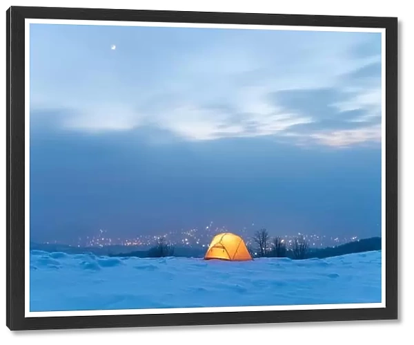 Yellow tent lighted from the inside against the backdrop of glowing city lights in fog. Amazing snowy landscape. Tourists camp in winter mountains
