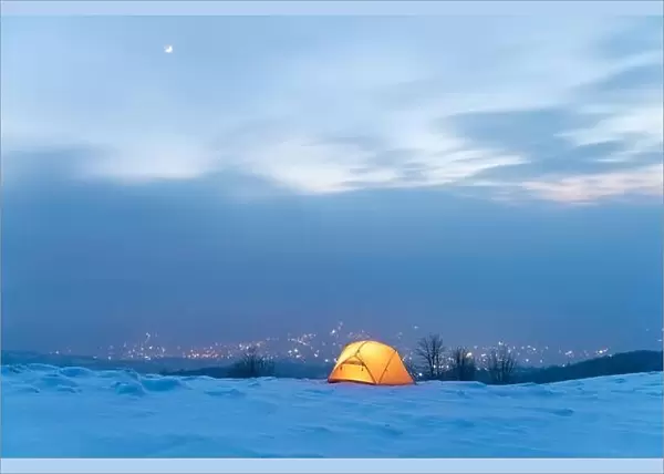 Yellow tent lighted from the inside against the backdrop of glowing city lights in fog. Amazing snowy landscape. Tourists camp in winter mountains