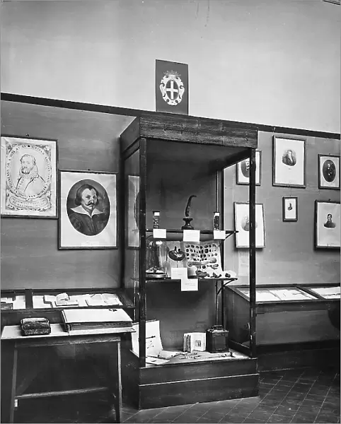 Room with some portraits hung on the walls, some books and other objects belonging to the University of Pavia. In the central glass case are some mounted animals, by Professor of Anthropology, Paolo Mantegazza, between 1862 and 1866, prepared by Rusconi. In the center is an exhibit on the galvanoplasty in 1800-1802 of doctor and chemist Luigi Brugnatelli. Some devices of Bottini are visible below. The photo was taken on the occasion of the Exhibition on the History of Science held in Florence in 1929