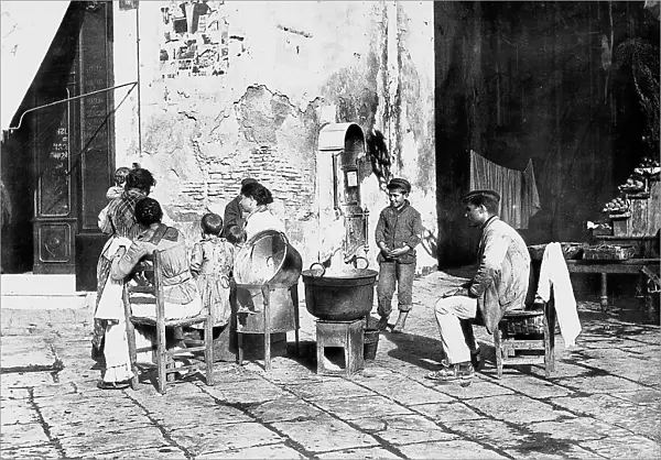 A group of men, women and children sitting in a piazza in Naples around some pots. On their right are some meats on a table and a staircase