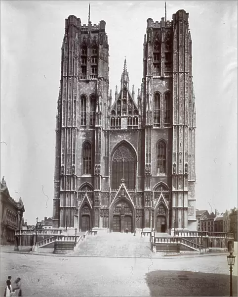 The facade of the Cathedral of Brussels dedicated to Saints Gudule and Michael. The church in gothic style has two large towers on the facade decorated with small spires and windows. In the foreground two passersby