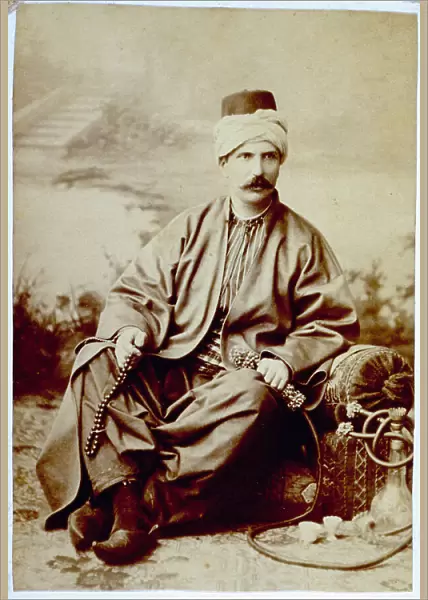 Portrait of a young european man in traditional turkish dress. He is seated on the ground and holds a koboloi in one hand and a water pipe in the other