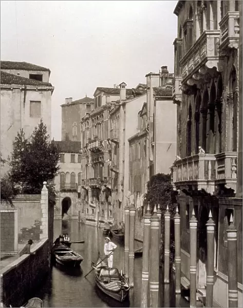 A venetian calle with a gondola going by. Other vessels are drawn up along the buildings. In the foreground a few mooring poles