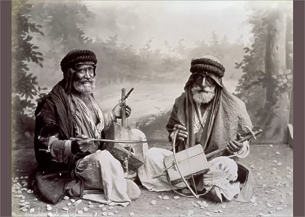 Portrait of two arab musicians in humble ethnic dress. The elderly men are posing in the studio, seated on the ground. Behind them is a backdrop of a wooded landscape