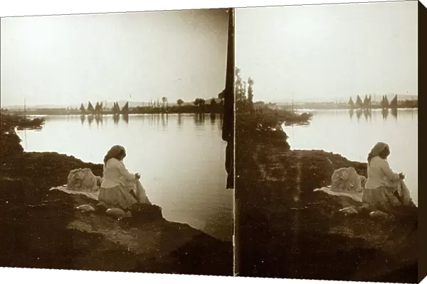 View from the banks of a river. In the foreground a woman in traditional attire is seated. In the background, sail boats