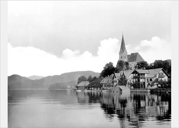 Group of houses among the trees along a bank of the Wrther See (Austria); among the buildings, a church can be seen. On the horizon are hills that surround the lake. The sky is partially covered by clouds