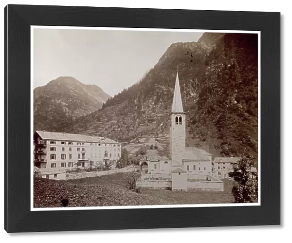 View of Alagna at the foot of the alpine mountain chain. Detail showing the church with its bell tower and spire, portico and cemetery, and a large hotel. The religious complex is surrounded by a lush vegetation