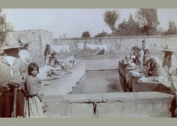 Mexican washerwomen at work around a tank of water; with them a few men and some little girls. In the background some clothes drying
