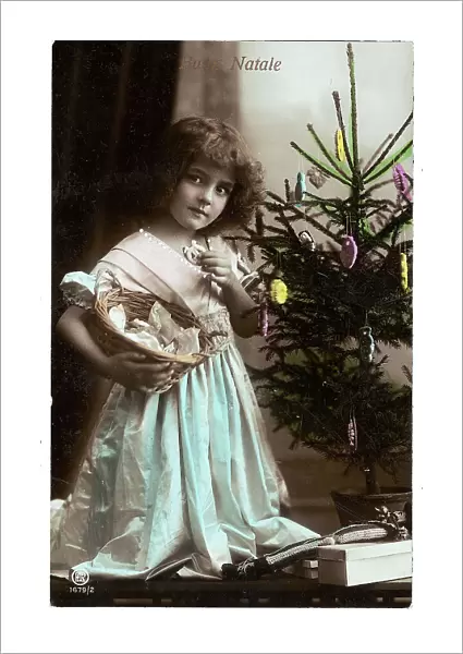 Portrait of a young little girl while eating a cookie and carrying a basket under the Christmas tree, Christmas greeting post-card, with a Buon Natale inscription on the front side and a personal dedication on the back side, the postage stamp indicates the date of 24th of December 1914, Peschiera, Verona, Italy