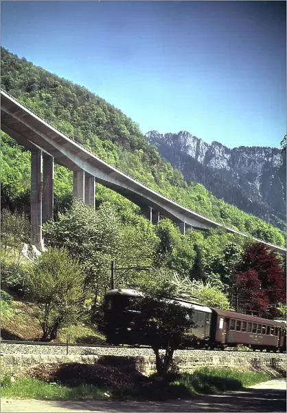 View of a mountain landscape crossed by a viaduct. At the bottom of the image, a train travels by on a railway