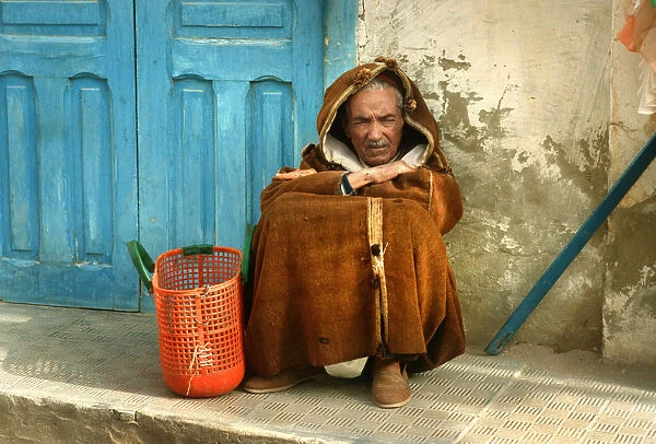 Old man wearing a djelleba sits on pavement, Tunisia