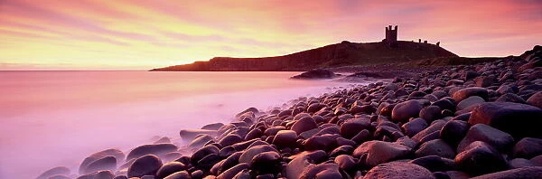 An imposing silhouette of Dunstanburgh Castle against a magnificent sky at sunrise with a beach of basalt boulders in the foreground, Embleton Bay, near Alnwick, Northumberland, England, United