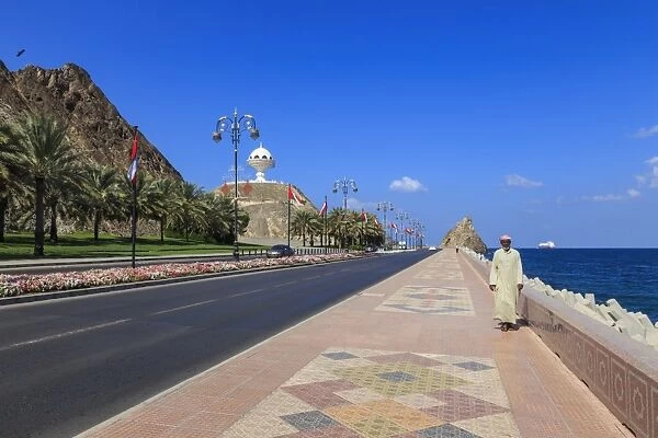 Man wearing dishdasha walks along Mutrah Corniche with national flags, flower beds