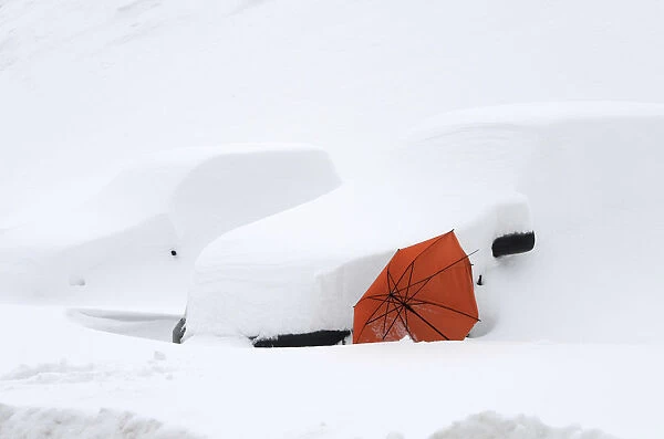 An abandoned red umbrella is supported by two cars covered in snow in the village of