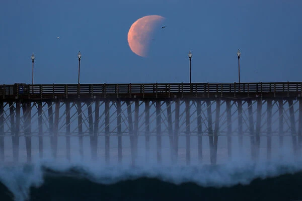 A Blue Moon comes out of a lunar eclipse as it sets past an ocean pier in Oceanside