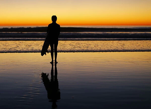 A lone surfer stands watching the waves after surfing as the sun sets in Leucadia