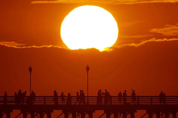 The sun sets behind a cloud as people cool off with a walk along an ocean pier in