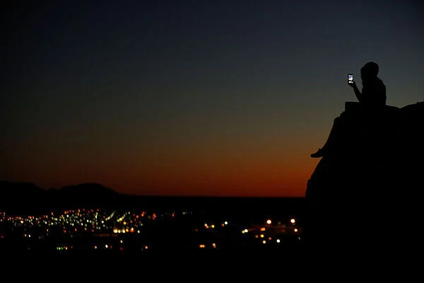 A young couple look at their phone as they sit on a hillside after sun set in El Paso