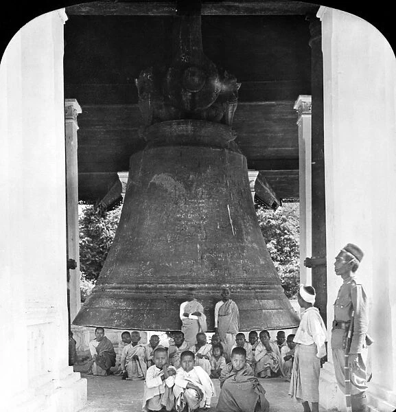 The Great Bell of Mingoon with a group of Buddhist monks posed in front of it and a guard standing to the right, at the temple in Mingoon, Burma. Stereograph, c1907