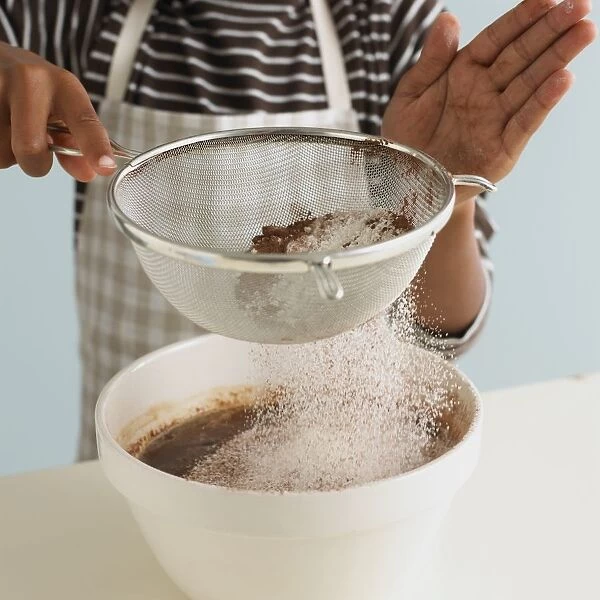 Boy sifting flour, cocoa, and baking powder onto chocolate mixture in mixing bowl (making chocolate brownies), close-up