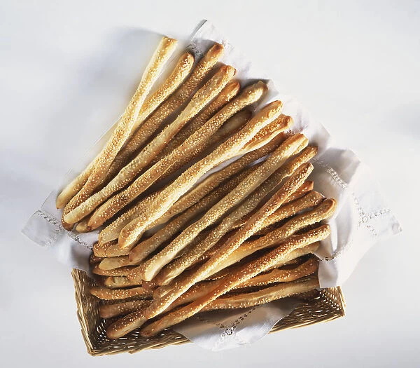 Breadsticks, sprinkled with sesame seeds, spilling out of wicker basket lined with white napkin, view from above