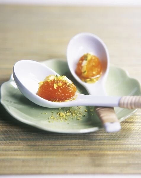 Two ceramic spoons containing dried apricots stuffed with cream and sprinkled with pistachios on wooden table, close-up