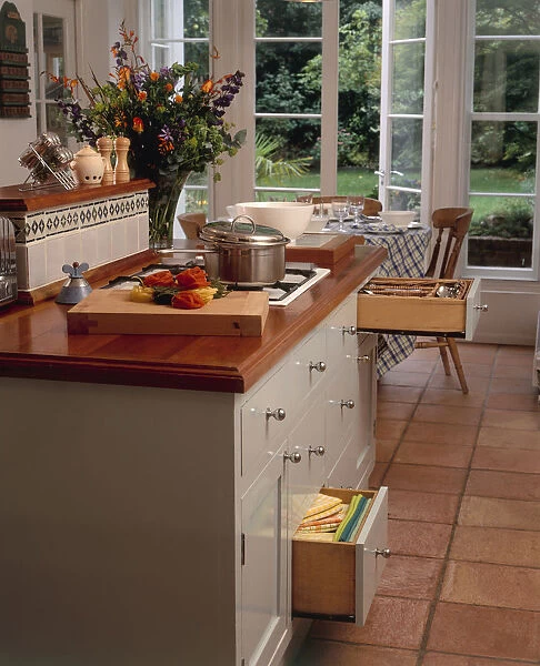Kitchen in white and brown tones, with terracotta tiled floor, drawer unit underneath hob, and dining table with door open onto garden