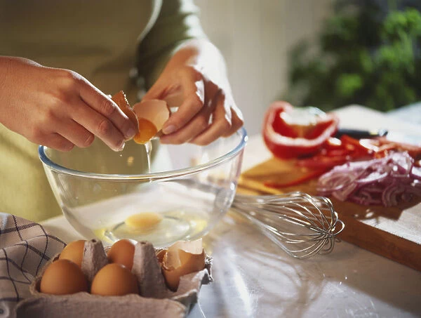 Raw egg dripping from shell being cracked over glass bowl, egg carton and whisk by sides of bowl, chopped vegetables on wooden board in background